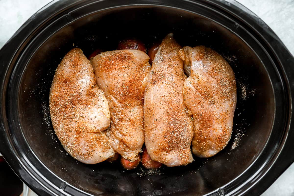 seasoned raw chicken breasts over red baby potatoes in a crockpot.