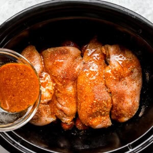 pouring sauce over seasoned raw chicken breasts and red baby potatoes in a crockpot.
