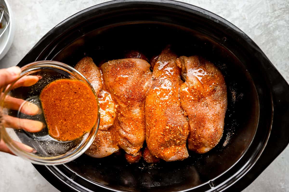 pouring sauce over seasoned raw chicken breasts and red baby potatoes in a crockpot.