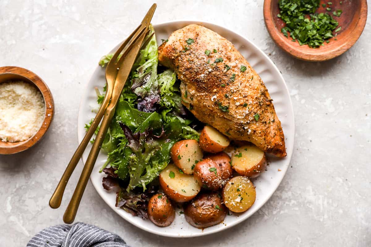 overhead view of a crockpot chicken breast and potatoes on a white plate with a side salad and cutlery.