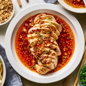 sliced mouthwatering chicken drenched in sauce on a white plate, surrounded by bowls of toppings.