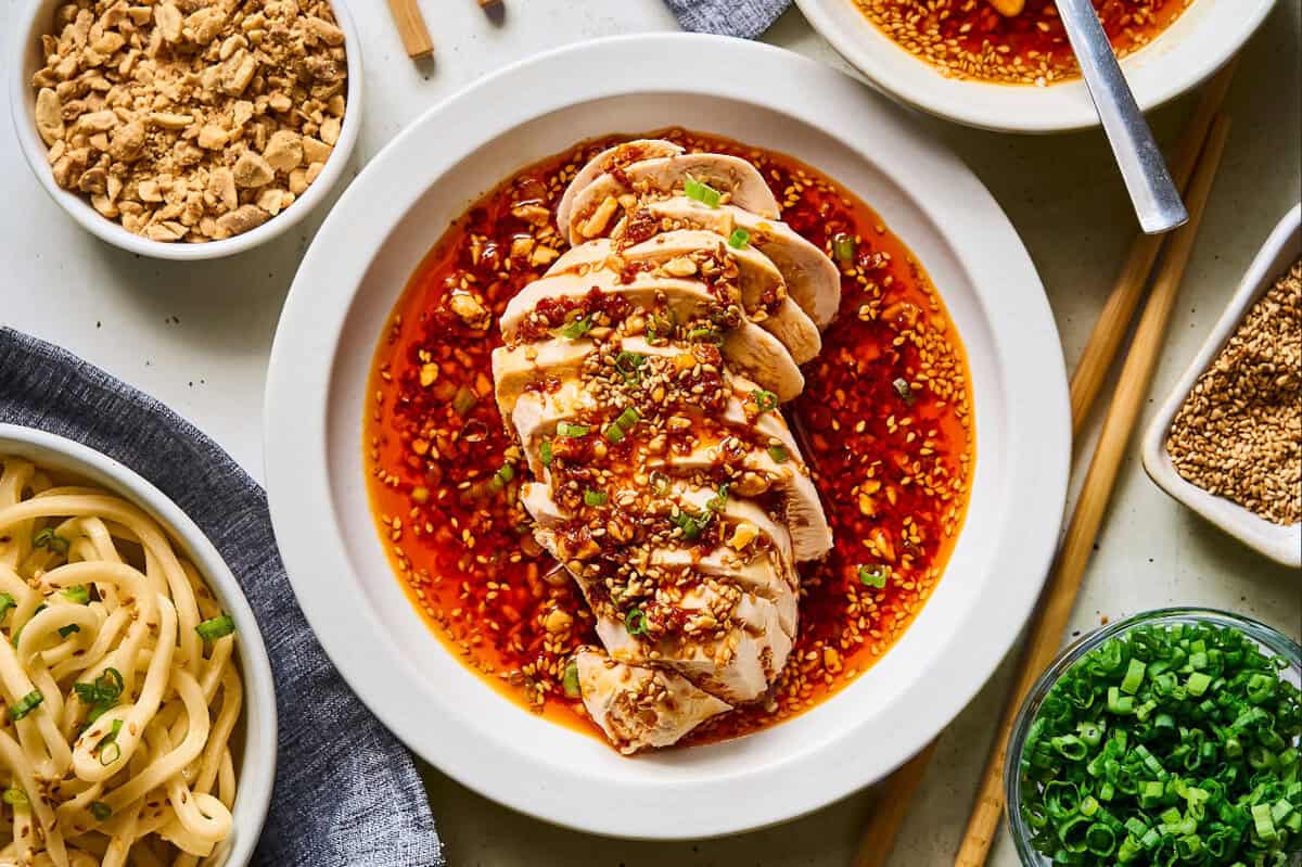 sliced mouthwatering chicken drenched in sauce on a white plate, surrounded by bowls of toppings.