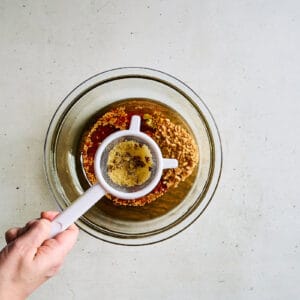 straining the solids from fragrant oil over spices in a bowl.