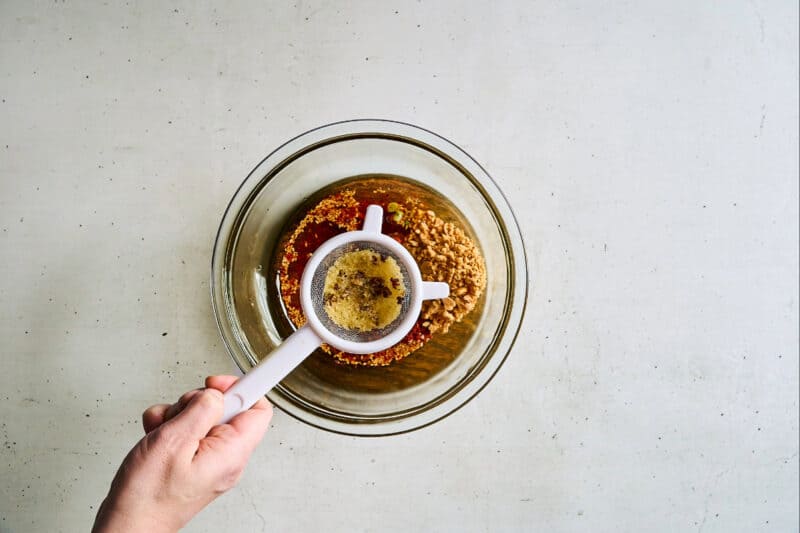 straining the solids from fragrant oil over spices in a bowl.