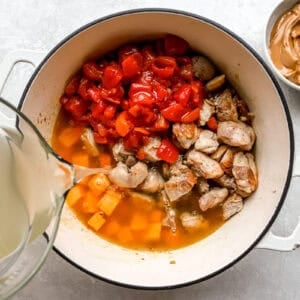 pouring broth into a pot with chicken, tomatoes, sweet potato and more.