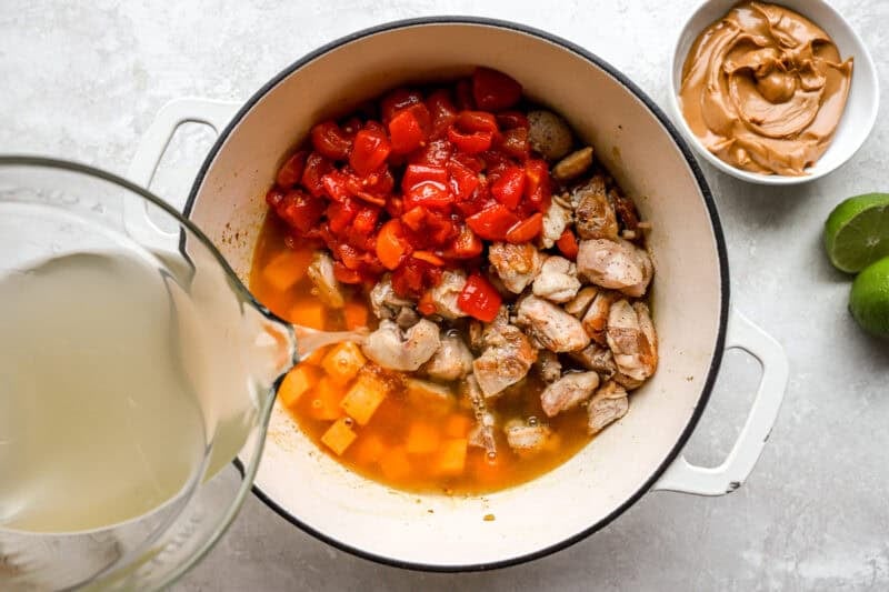 pouring broth into a pot with chicken, tomatoes, sweet potato and more.
