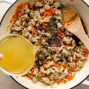 pouring chicken broth over seasoned ground chicken and vegetables in a dutch oven.
