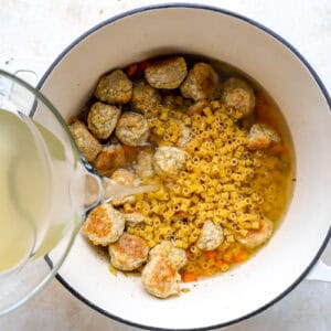 pouring chicken broth over mirepoix, chicken meatballs, and ditalini pasta in a Dutch oven.