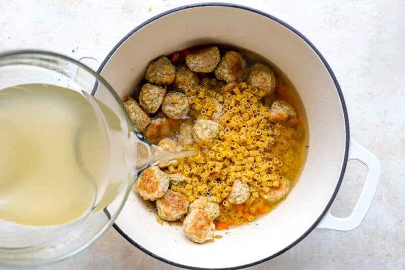 pouring chicken broth over mirepoix, chicken meatballs, and ditalini pasta in a Dutch oven.