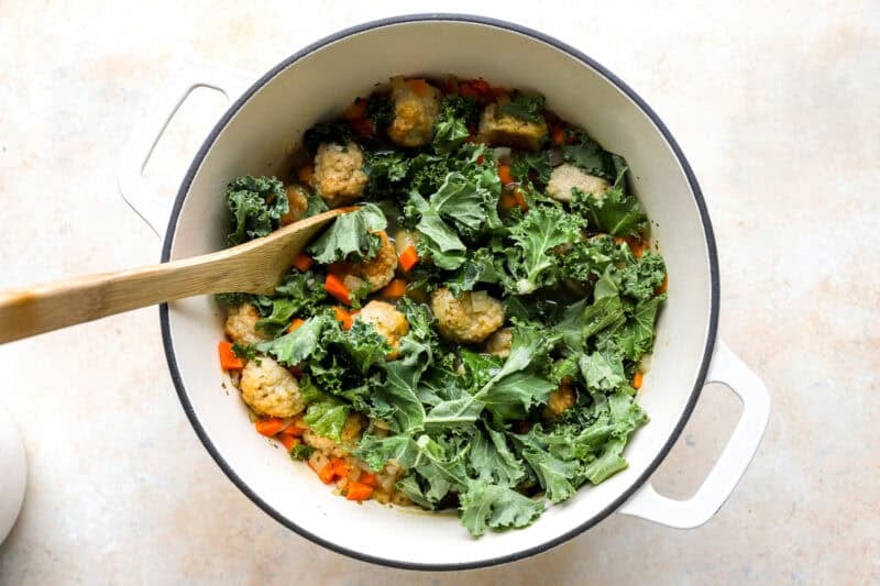 stirring kale into chicken meatball soup in a Dutch oven with a wooden spoon.