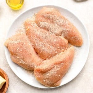 four seasoned raw chicken breasts on a white plate.