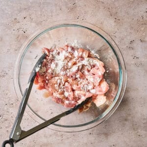 sliced chicken thighs tossed with cornstarch in a glass bowl with tongs.