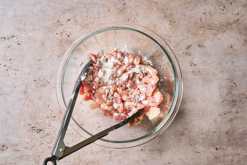 sliced chicken thighs tossed with cornstarch in a glass bowl with tongs.