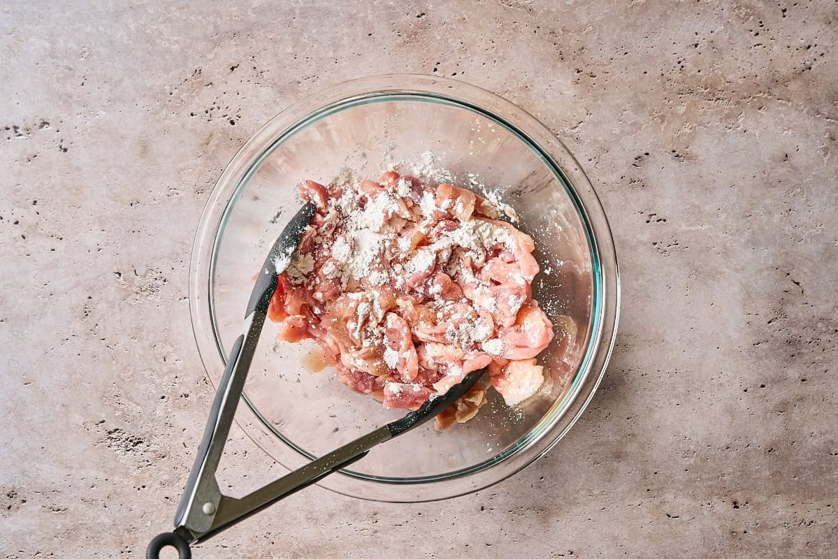 sliced chicken thighs tossed with cornstarch in a glass bowl with tongs.