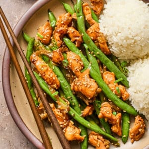 overhead view of a serving of chicken and green bean stir fry with sesame seeds and a side of white rice on a plate.