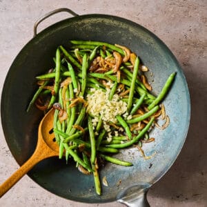 adding garlic to stir fried green beans and onions in a wok with a wooden spoon.