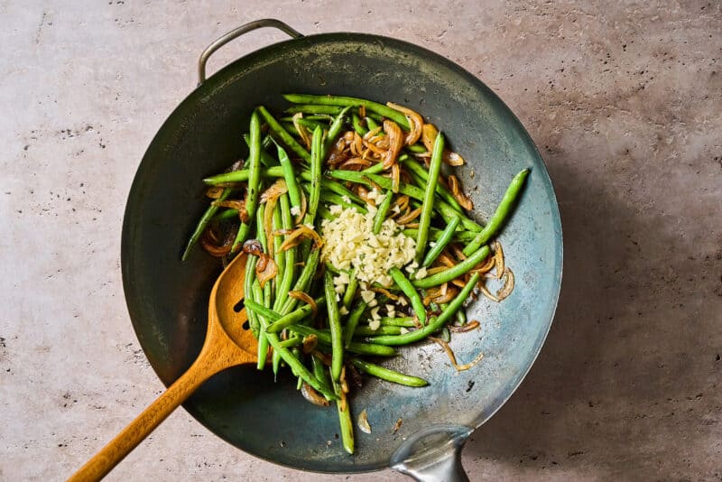 adding garlic to stir fried green beans and onions in a wok with a wooden spoon.