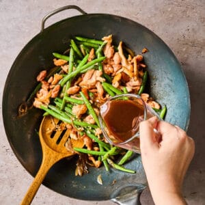 pouring hoisin sauce over chicken and green bean stir fry in a wok with a wooden spoon.