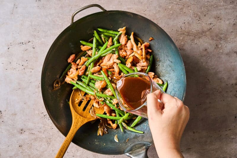 pouring hoisin sauce over chicken and green bean stir fry in a wok with a wooden spoon.