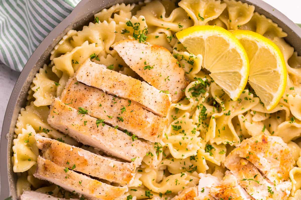 Sliced lemon chicken breasts served in a bowl of pasta with lemon butter sauce, lemon wedges, and parsley.