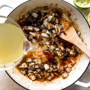 pouring chicken broth over seasoned onions and chili peppers in a pan with a wooden spoon.