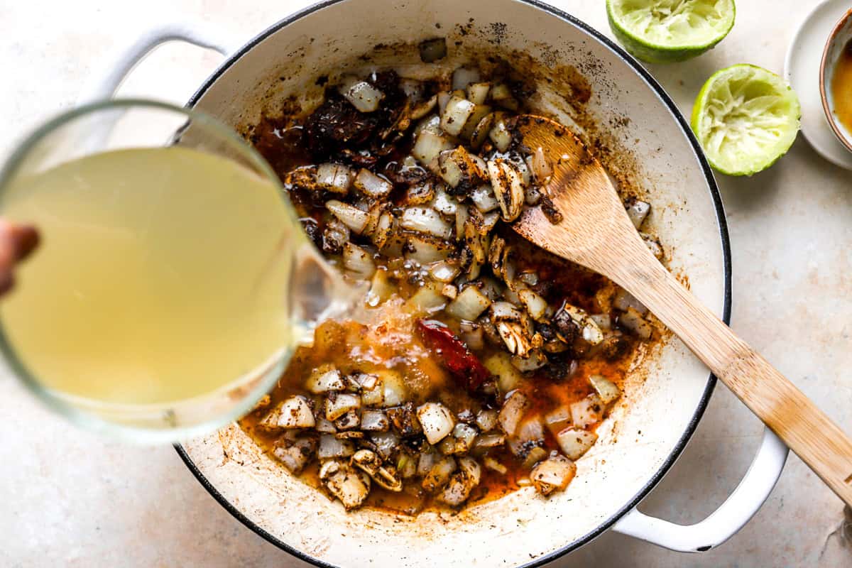 pouring chicken broth over seasoned onions and chili peppers in a pan with a wooden spoon.