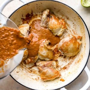 pouring blended birria sauce over seared chicken thighs in a pan.