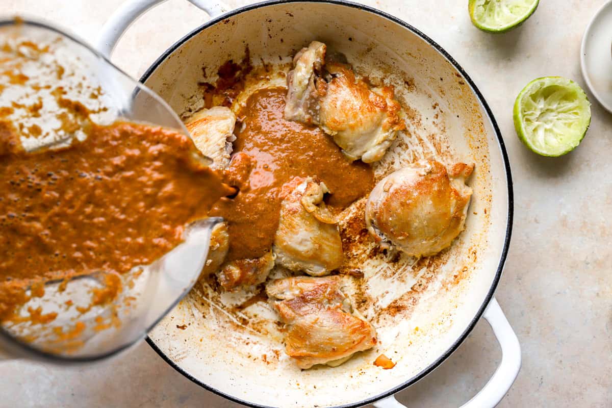 pouring blended birria sauce over seared chicken thighs in a pan.