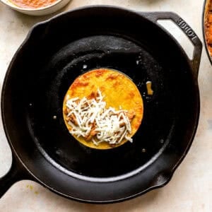 a small corn tortilla topped with chicken birria and cheese in a cast iron skillet.