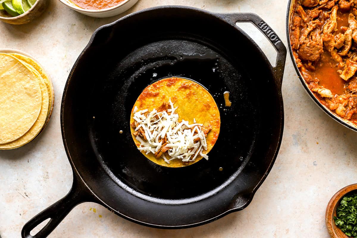 a small corn tortilla topped with chicken birria and cheese in a cast iron skillet.