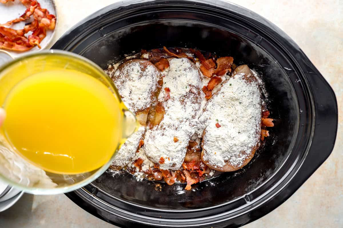 pouring chicken broth over seasoned chicken breasts and veggies in a crockpot.