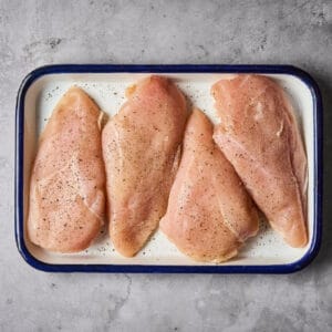four seasoned raw chicken breasts on a baking pan.