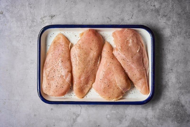 four seasoned raw chicken breasts on a baking pan.