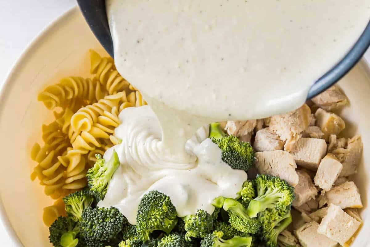 Pouring Alfredo sauce into a bowl of cubes chicken, broccoli, and pasta.