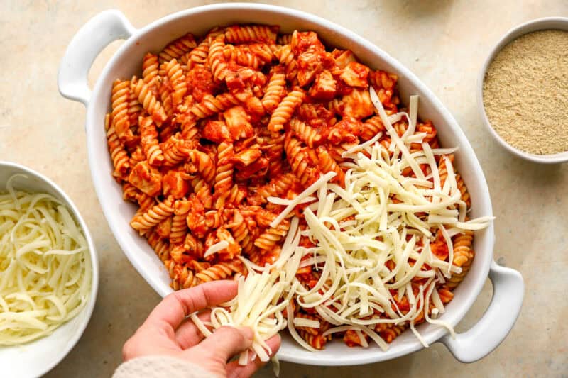 sprinkling shredded mozzarella cheese over chicken parmesan casserole in a baking dish.