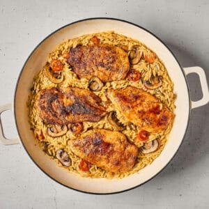 vegetable orzo topped with seared chicken breasts in a pan.