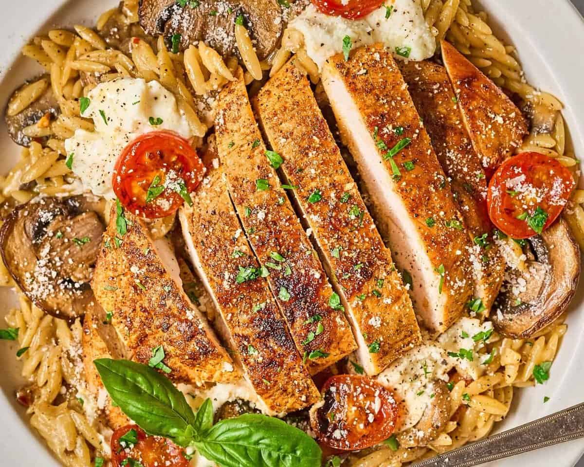 closeup view of a sliced chicken breast over vegetable orzo and ricotta cheese with a fork.