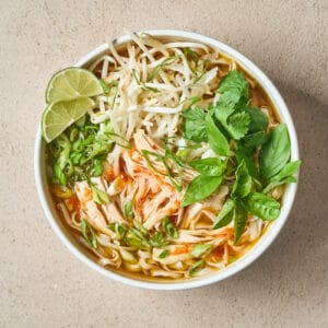 a bowl of chicken pho with bean sprouts, fresh greens, and lime slices.