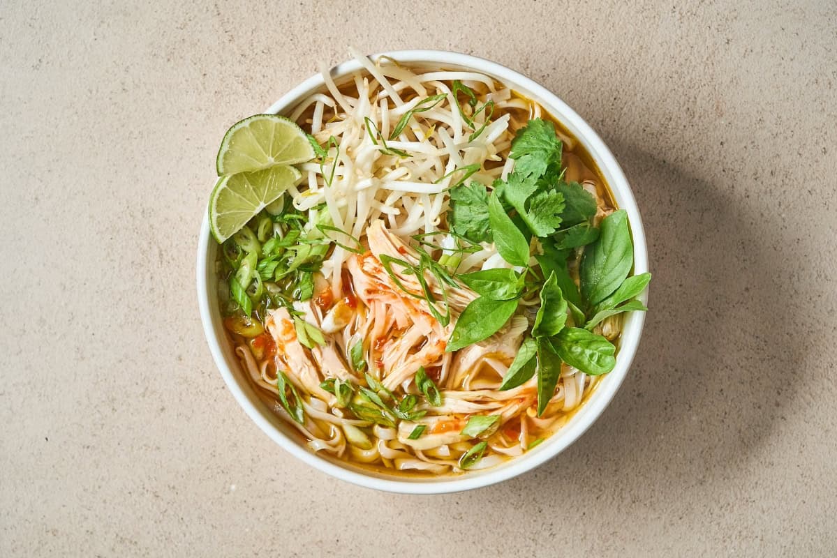 a bowl of chicken pho with bean sprouts, fresh greens, and lime slices.