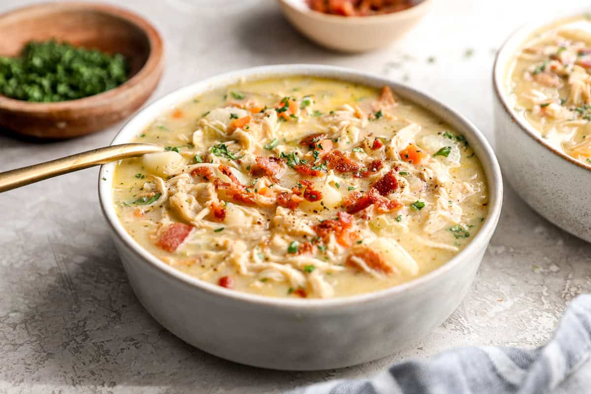 three-quarters view of a bowl of creamy chicken and potato soup with a spoon, topped with bacon and parsley.