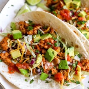 closeup of ground chicken tacos on a white plate topped with chopped avocado, red onion, and salsa.