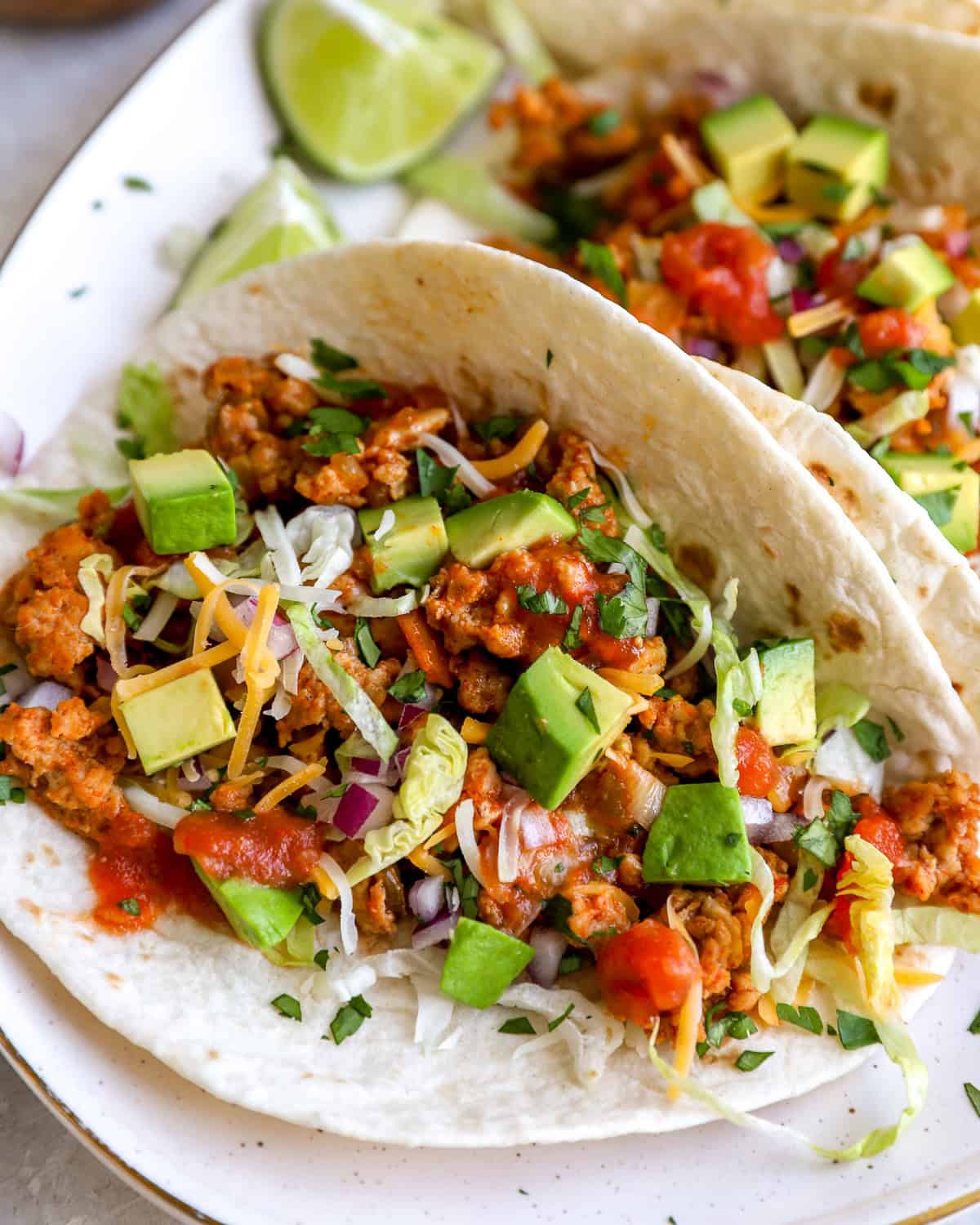 closeup of ground chicken tacos on a white plate topped with chopped avocado, red onion, and salsa.