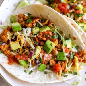 closeup of ground chicken tacos on a white plate topped with chopped avocado, red onion, and salsa.