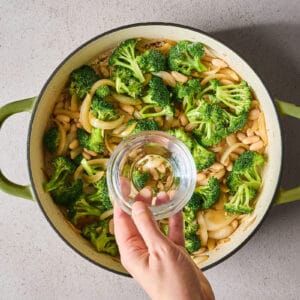 pouring a small amount of water over broccoli, white beans, onions, and garlic in a skillet.