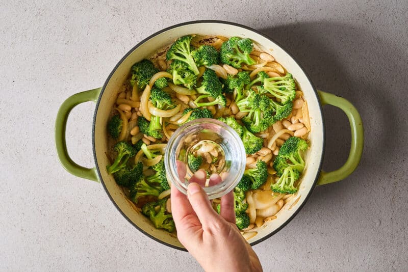pouring a small amount of water over broccoli, white beans, onions, and garlic in a skillet.