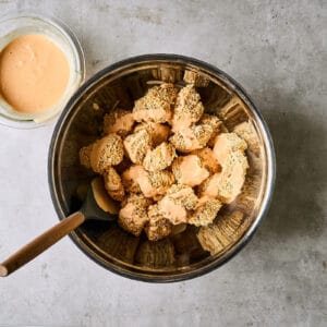 tossing baked panko-coated chicken pieces in bang bang sauce in a metal bowl with a rubber spatula.