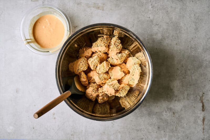 tossing baked panko-coated chicken pieces in bang bang sauce in a metal bowl with a rubber spatula.