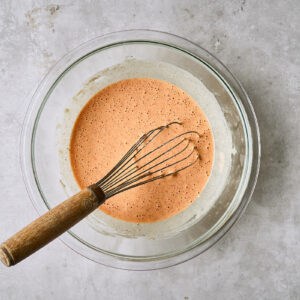 orange-colored batter in a glass bowl with a whisk.