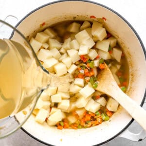 pouring chicken broth over sautéed veggies and diced potatoes in a dutch oven.