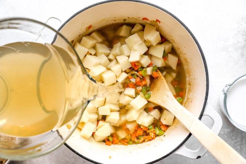 pouring chicken broth over sautéed veggies and diced potatoes in a dutch oven.